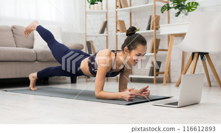 A woman in athletic wear is doing a plank exercise on a yoga mat while watching a laptop. She is in her living room with a couch and bookshelf in the background. 116612893