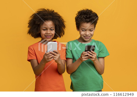 Two African American children, a girl and a boy, are standing in front of a yellow background, smiling while looking down at their smartphones. Two African American children, a girl and a boy, are standing in front of a yellow background, smiling while looking down at their smartphones. 116612949