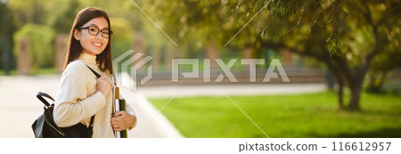 A young Chinese woman in a white sweater and glasses walks through a park. She is carrying a backpack and holding a stack of books in her left hand, panorama with copy space 116612957