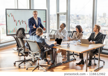 A business leader presents financial data to his team during a meeting in a modern office. Leader stands near a large screen displaying a chart, while the team members sit at a table in front of him 116612982