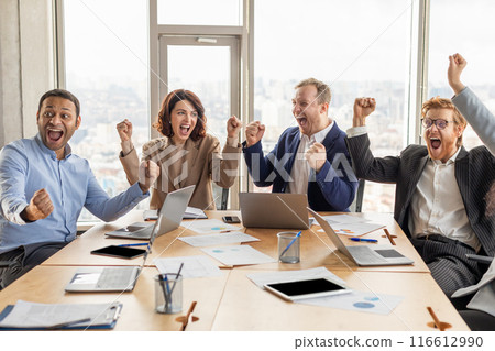 A diverse team of four business professionals are sitting at a conference table in a modern office, all smiling and raising their arms in celebration. 116612990