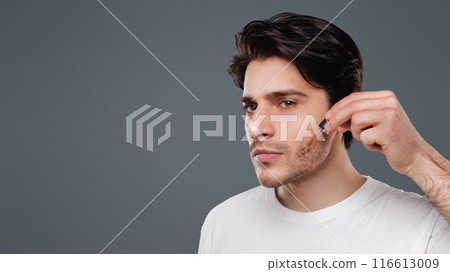 Handsome man applying moisturizing serum on face, grey studio background 116613009