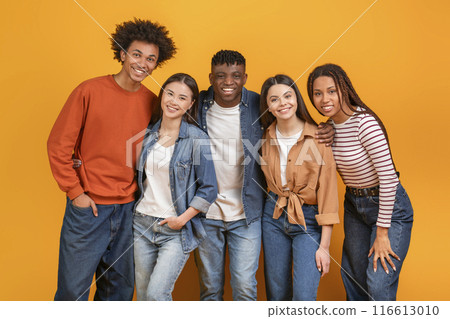 Group of five diverse young adults standing in front of a yellow background. They are all smiling and looking at the camera. The group appears to be friends and is enjoying each others company. 116613010