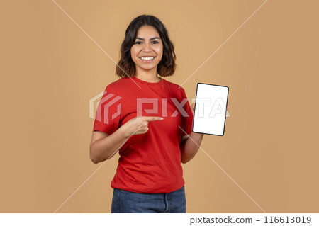 A young middle eastern woman with a bright smile is holding a tablet with a blank white screen. She is wearing a red t-shirt and blue jeans. The background is a light brown color. 116613019