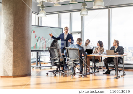 A businessman in a suit stands in front of a whiteboard displaying a financial chart, presenting to a small group of colleagues seated around a table in a modern office with large windows. A businessman in a suit stands in front of a whiteboard displaying a financial chart, presenting to a small group of colleagues seated around a table in a modern office with large windows. 116613029