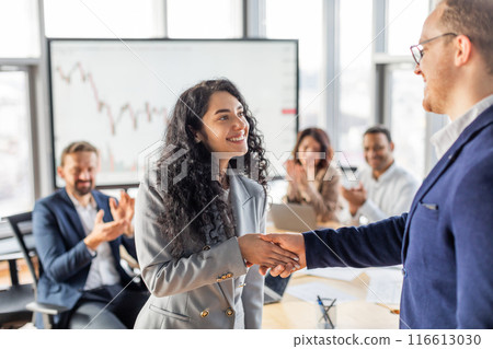 A businesswoman in a grey blazer shakes hands with a colleague wearing a blue suit. Other colleagues are clapping in the background, indicating a successful presentation or achievement. A businesswoman in a grey blazer shakes hands with a colleague wearing a blue suit. Other colleagues are clapping in the background, indicating a successful presentation or achievement. 116613030
