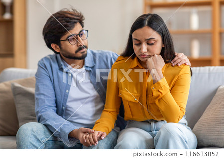 Indian man with glasses and a blue shirt sits on a couch beside a woman in a yellow top, comforting her as she appears upset. He holds her hand and gently rests his other hand on her shoulder Indian man with glasses and a blue shirt sits on a couch beside a woman in a yellow top, comforting her as she appears upset. He holds her hand and gently rests his other hand on her shoulder 116613052