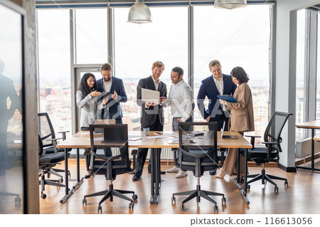A group of business professionals are having a meeting in a modern office. They are standing around a large wooden table, looking at laptops and tablets, and discussing work. A group of business professionals are having a meeting in a modern office. They are standing around a large wooden table, looking at laptops and tablets, and discussing work. 116613056