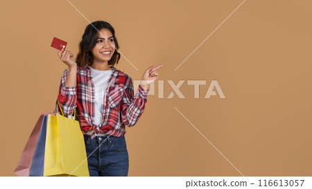 A young arab woman with short, wavy brown hair smiles at the camera while holding a credit card and several shopping bags, pointing at copy space A young arab woman with short, wavy brown hair smiles at the camera while holding a credit card and several shopping bags, pointing at copy space 116613057