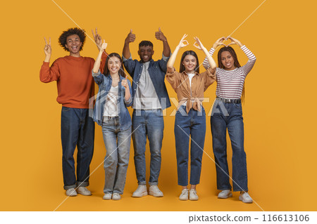 Multiethnic group of young adults, stand together in front of a solid yellow background. They are all smiling and making various hand gestures, including peace signs, thumbs up, and a heart shape Multiethnic group of young adults, stand together in front of a solid yellow background. They are all smiling and making various hand gestures, including peace signs, thumbs up, and a heart shape 116613106