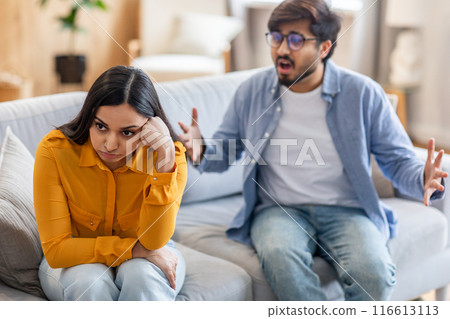 A young Indian couple is sitting on a couch in their living room engaged in a heated argument. The woman appears upset and is looking away, while the man is gesturing passionately with his hands. A young Indian couple is sitting on a couch in their living room engaged in a heated argument. The woman appears upset and is looking away, while the man is gesturing passionately with his hands. 116613113