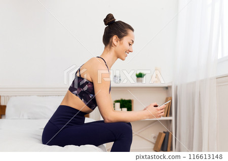 A woman in activewear sits on the edge of a bed, looking at her phone. A woman in activewear sits on the edge of a bed, looking at her phone. 116613148