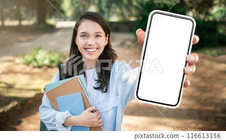 A young lady student smiles brightly as she holds out a smartphone with a blank screen in front of her. She is standing outdoors in a park-like setting A young lady student smiles brightly as she holds out a smartphone with a blank screen in front of her. She is standing outdoors in a park-like setting 116613156