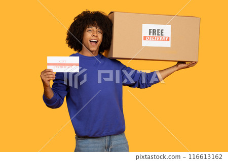 A young man with curly hair wearing a blue sweater holds up a gift certificate and a delivery box labeled Free Delivery. He appears delighted, celebrating a surprise or special delivery 116613162