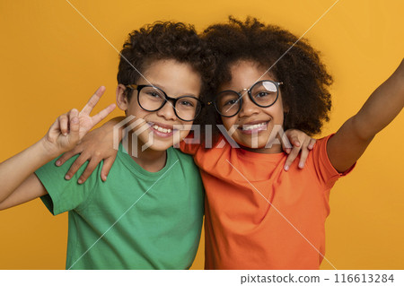Two young African American children, a boy and a girl, posing for a photograph. The boy is wearing a green t-shirt and glasses, while the girl is wearing an orange t-shirt and glasses. 116613284