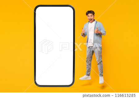 A young man jumps in the air in front of a large smartphone with a blank white screen, giving a thumbs up with both hands and a big smile on his face. The background is a bright yellow 116613285