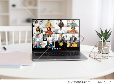 A laptop on a white table displays a virtual team meeting with multiple participants. The bright, modern home office background includes a white bookshelf, green plant, and geometric decor piece. 116613296