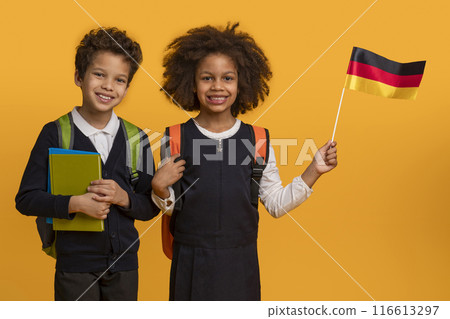 Two black children, a boy and a girl, are standing in front of a yellow background. They are both wearing school uniforms and smiling at the camera. The girl is holding a German flag Two black children, a boy and a girl, are standing in front of a yellow background. They are both wearing school uniforms and smiling at the camera. The girl is holding a German flag 116613297