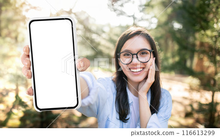 A young woman with long dark hair, wearing glasses and a light blue shirt, smiles brightly as she holds a smartphone with a blank white screen out in front of her A young woman with long dark hair, wearing glasses and a light blue shirt, smiles brightly as she holds a smartphone with a blank white screen out in front of her 116613299