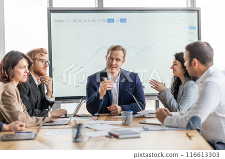 A group of business professionals are seated around a conference table, engaged in a discussion. They are gathered in a modern office with a large whiteboard behind them 116613303