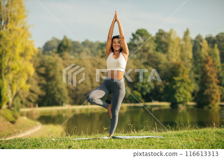 A woman is practicing yoga outside near a tranquil lake. She is focused and engaged in various yoga poses, surrounded by nature and fresh air. 116613313