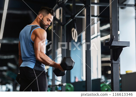 A man in a gym setting lifts a dumbbell during a workout. He is wearing a sleeveless shirt and black pants. A man in a gym setting lifts a dumbbell during a workout. He is wearing a sleeveless shirt and black pants. 116613346