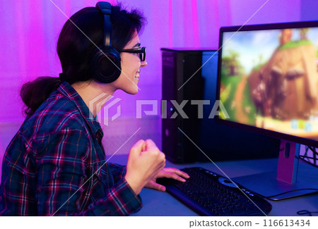 A young woman is pictured playing a video game on a desktop computer. She is wearing a blue and red plaid shirt and headphones and is excitedly pumping her fist in the air. A young woman is pictured playing a video game on a desktop computer. She is wearing a blue and red plaid shirt and headphones and is excitedly pumping her fist in the air. 116613434