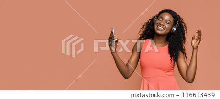 A young black woman with long curly hair is dancing and listening to music on headphones while holding a smartphone in her hand. She is smiling and enjoying the music. A young black woman with long curly hair is dancing and listening to music on headphones while holding a smartphone in her hand. She is smiling and enjoying the music. 116613439