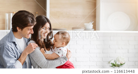 This image shows a happy family of three in a modern kitchen. The parents are smiling as they hold their baby, who is looking off to the side, copy space 116613448
