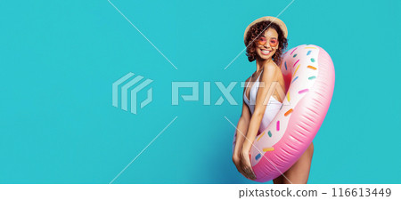 A young black woman in a white swimsuit and a straw hat smiles while holding a pink donut-shaped inflatable pool float in front of a bright blue background, copy space A young black woman in a white swimsuit and a straw hat smiles while holding a pink donut-shaped inflatable pool float in front of a bright blue background, copy space 116613449