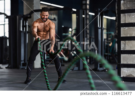 Crossfit training. Naked muscular man exercising with battle ropes at gym, free space Crossfit training. Naked muscular man exercising with battle ropes at gym, free space 116613477