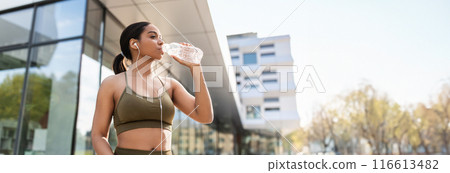 Young black woman in sportswear drinking water, listening to music in earphones during training on city street. Fit millennial African American lady keeping hydrated while jogging outdoors, copy space Young black woman in sportswear drinking water, listening to music in earphones during training on city street. Fit millennial African American lady keeping hydrated while jogging outdoors, copy space 116613482