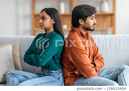 A young Indian couple is sitting back-to-back on a couch, clearly upset and avoiding eye contact. Both are crossing their arms, conveying signs of frustration and disagreement. 116613484
