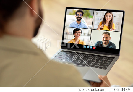 A man sitting at a table is using a laptop, which displays a group of people on the screen. He appears engaged in a virtual meeting or video conference with his colleagues. 116613489