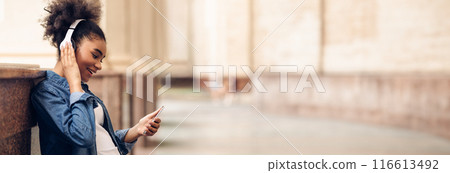 Black Student Girl Using Smartphone Wearing Headphones Listening To Music Or Educational Podcast Standing Outdoor Near College Building. People And Gadgets, Side View, Copy Space Black Student Girl Using Smartphone Wearing Headphones Listening To Music Or Educational Podcast Standing Outdoor Near College Building. People And Gadgets, Side View, Copy Space 116613492