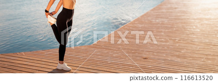 Unrecognizable sporty lady stretching legs, warming up before jogging outdoors, cropped shot of athletic woman in activewear exercising on wooden pier near water, enjoying training outside, copy space 116613520