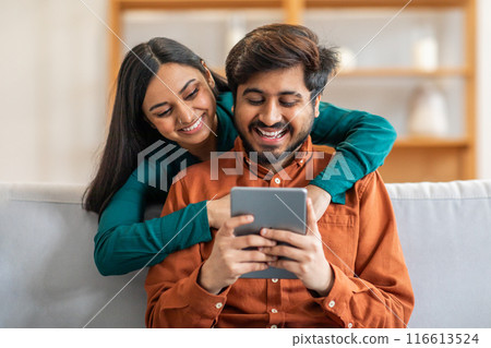 Indian man and woman are seated on a couch, both focused on a tablet screen. They appear engaged as they interact with the device, possibly watching a video or scrolling through information 116613524