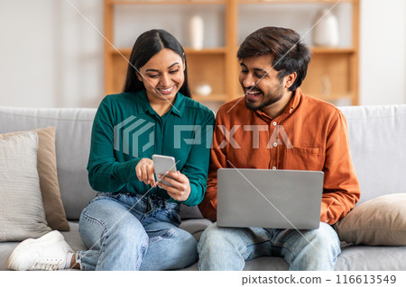 A smiling Indian couple is seated on a comfortable sofa in their modern living room, enjoying leisure time together. The woman is looking at her smartphone while the man is using a laptop 116613549