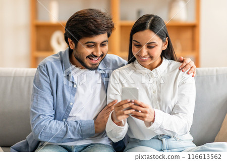 Indian couple sitting on a couch, both focused on looking at a cell phone screen. They appear engaged in whatever is displayed on the device, with expressions of concentration and interest. 116613562