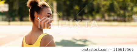 Portrait of half-turned smiling African American lady wearing wireless headphones, exercising outdoors. Woman runner listening to music with ear buds after running outside, copy space 116613566