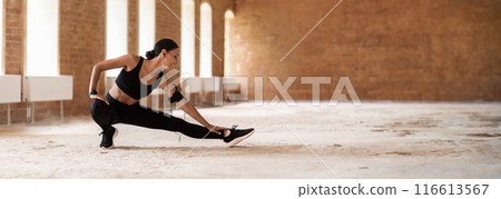 Smiling young slim arab lady in sportswear with phone and fitness tracker in headphones doing leg exercises in loft building interior, panorama with copy space 116613567