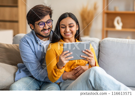 Indian man and woman are seated on a couch, both focused on a tablet in front of them. They seem engaged in the digital device, possibly discussing or sharing information. 116613571