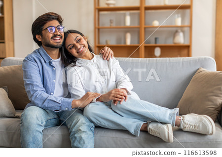 In the living room, Indian man and woman are seated on a couch. They appear relaxed and engaged in conversation, facing each other. The room is well-lit, and the decor is simple and modern. 116613598