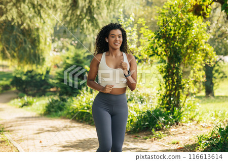 A woman in sportswear is jogging along a paved path in a park. The path is lined with trees, and the surroundings are green and lush. 116613614