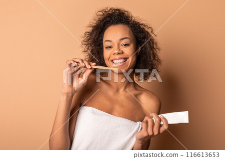 African American young woman with curly hair smiles brightly while brushing her teeth with a bamboo toothbrush. She is wrapped in a white towel and holds a tube of toothpaste in her other hand African American young woman with curly hair smiles brightly while brushing her teeth with a bamboo toothbrush. She is wrapped in a white towel and holds a tube of toothpaste in her other hand 116613653