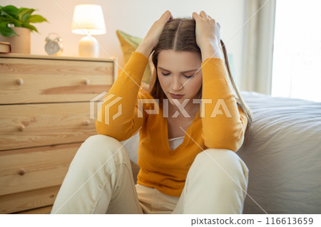A teen girl, dressed in a yellow sweater and light pants, sits on her bedroom floor looking distressed with her hands on her head. Daylight illuminates the room from a nearby window. 116613659