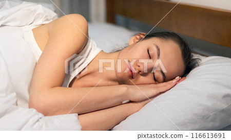 A young woman is sleeping soundly in bed. She is lying on her side with her head resting on a pillow. 116613691