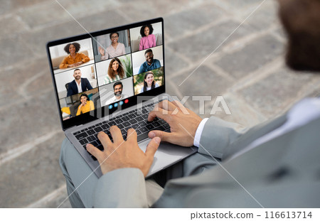 African American business professional is sitting outdoors, engaged in a virtual meeting with multiple colleagues displayed on a laptop screen. The person is typing on the keyboard African American business professional is sitting outdoors, engaged in a virtual meeting with multiple colleagues displayed on a laptop screen. The person is typing on the keyboard 116613714
