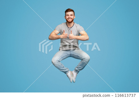 Enjoy life moments. Joyful redhaired man jumping in air, demonstrating thumbs up gesture, blue studio background, full length, free space 116613737