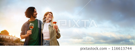 A young couple stands together on a beach, enjoying a glass of wine as they watch the sunset over the water. They are both smiling and looking at each other, panorama with copy space 116613744
